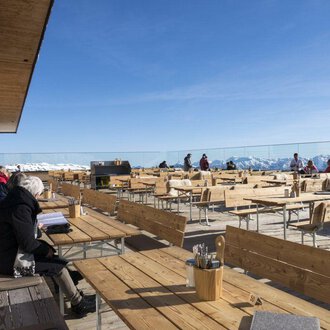 An idyllic restaurant on a mountain terrace with a variety of wooden tables and chairs. In the background, snow-capped mountains and a clear sky can be seen. | © OBERSTDORF · KLEINWALSERTAL BERGBAHNEN