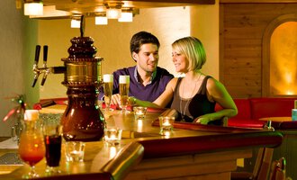 Ein gemütlicher Barbereich mit einem Paar, das sich unterhält. Im Hintergrund sind verschiedene Getränke und eine warme Holzdekoration zu sehen. | © Aparthotel Kleinwalsertal Haller GmbH & Co KG