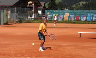 A man is playing tennis on a red court with a view of the mountains. In the background, other players and a tennis wall can be seen. | © Sportverein Casino Kleinwalsertal