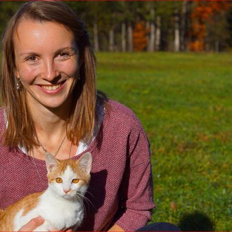 A smiling woman is sitting on a meadow holding an orange-and-white cat. Autumn trees can be seen in the background. | © Tierarztpraxis Oberstdorf | Regina Baumgartner