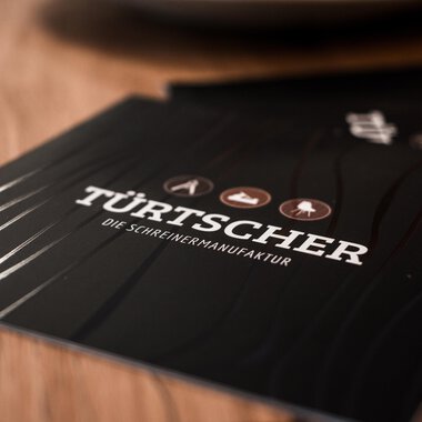 An elegant menu booklet with the inscription "TÜRTSCHER – the joiner's workshop". It features various wood symbols on a dark background. | © Türtscher Die Schreinermanufaktur