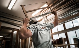 A craftsman holds a chair above his head and works in a workshop. In the background, wood materials and tools are visible. | © Türtscher Die Schreinermanufaktur