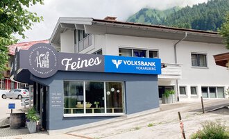A modern building with a business sign "Feines" and the Volksbank Vorarlberg. In the background, green hills and a clear sky can be seen. | © Volksbank Vorarlberg e. Gen.