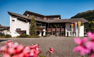 A modern building with an elegant facade, surrounded by beautiful flowers. In the background, mountains and a clear sky are visible. | © Walser Raiffeisen Bank AG