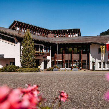 A modern building with an elegant facade, surrounded by beautiful flowers. In the background, mountains and a clear sky are visible. | © Walser Raiffeisen Bank AG