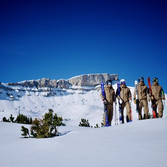 Eine Gruppe von Skifahrern steht auf einer schneebedeckten Fläche. Im Hintergrund sieht man beeindruckende Berggipfel und einen klaren blauen Himmel. | © Walser Skischule | Herbert Jochum