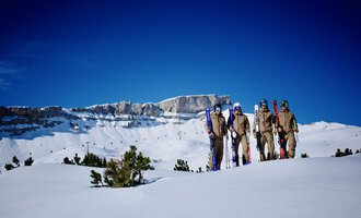 Eine Gruppe von Skifahrern steht auf einer schneebedeckten Fläche. Im Hintergrund sieht man beeindruckende Berggipfel und einen klaren blauen Himmel. | © Walser Skischule | Herbert Jochum
