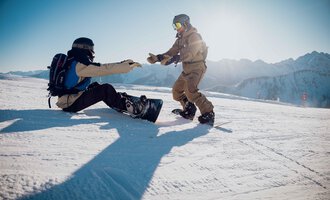 Zwei Snowboarder auf einer verschneiten Piste. Einer hilft dem anderen, der auf dem Boden sitzt. | © Walser Skischule | Ole Zumpolle