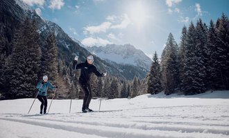 Zwei Personen beim Langlaufen in einer verschneiten Landschaft. Im Hintergrund sind majestätische Berge und eine klare, sonnige Himmel zu sehen. | © Walser Skischule | Ole Zumpolle