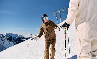 Ein Skifahrer mit einer braunen Skijacke steht auf einem schneebedeckten Hang. Im Hintergrund sind verschneite Berge und ein blauer Himmel zu sehen. | © Walser Skischule | Oliver Farys