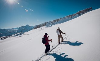 Zwei Wanderer gehen durch tiefen Schnee in den Bergen. Die Sonne scheint klar am blauen Himmel. | © Walser Skischule | Ole Zumpolle