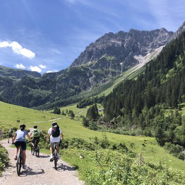 Eine Gruppe von Radfahrern fährt durch eine malerische Berglandschaft. Die grünen Wiesen und die majestätischen Berge schaffen eine wunderschöne Atmosphäre. | © WalserBike Tours | Christian Gutermuth