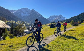 © WalserBike Tours | Christian Gutermuth Drei Radfahrer fahren auf einem Weg durch eine grüne Landschaft mit Bergen im Hintergrund. Der Himmel ist klar und blau. | © WalserBike Tours | Christian Gutermuth