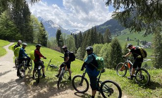 © WalserBike Tours | Christian Gutermuth Eine Gruppe von Radfahrern steht auf einem Weg in den Bergen. Die Umgebung ist grün mit Wald und einer beeindruckenden Berglandschaft im Hintergrund. | © WalserBike Tours | Christian Gutermuth