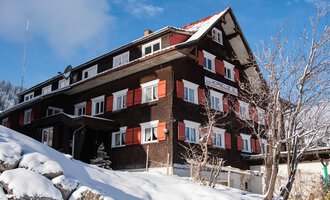 A large, traditional chalet in the mountains, surrounded by snow. The windows are decorated with red shutters, and the sky is clear and blue.