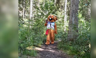 © Wildnisschule Kleinwalsertal Ein Maskottchen in einem Wald trägt eine Trikotnummer. Es steht auf einem schmalen Pfad zwischen den Bäumen. | © Wildnisschule Kleinwalsertal