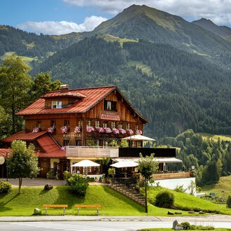 Ein malerisches Chalet in den Bergen mit blühenden Fensterkästen. Im Hintergrund erheben sich grüne Hügel und majestätische Berge.