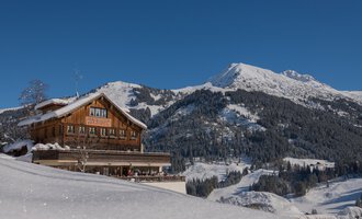 Ein wunderschönes Chalet im Schnee mit einem beeindruckenden Berg im Hintergrund. Der Himmel ist klar und blau, was die Winterlandschaft noch malerischer macht. | © Wirtshaus Hoheneck | Klaus Ruland