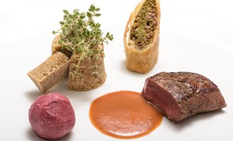 Ein kunstvoll angerichteter Teller mit saftigem Fleisch, einer grünen Kräuterdeko und verschiedenen Beilagen. Die Speisen sind ansprechend kombiniert und haben lebendige Farben. | © Wirtshaus Hoheneck | Klaus Ruland