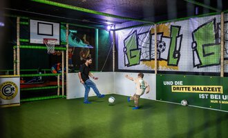 © Wonniland Sonthofen | interSPA Gruppe Ein spielender Vater und sein Kind in einer aufregenden Indoor-Sportarena. Sie kicken mit einem Fußball auf grünem Kunstrasen unter bunten Grafiken. | © Wonniland Sonthofen | interSPA Gruppe