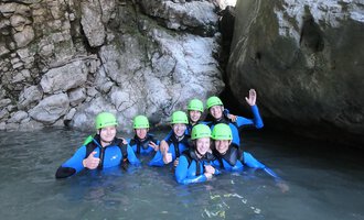 © Rocksolid Canyoning Tours Eine Gruppe von acht Personen in Neoprenanzügen und grünen Helmen steht im Wasser zwischen großen Felsen. Sie lächeln und zeigen Daumen hoch. | © Rocksolid Canyoning Tours