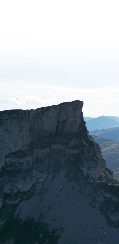 Ein dramatischer Berggipfel, der steil gegen einen hellen Himmel aufragt, mit entfernten Hügeln im Hintergrund.